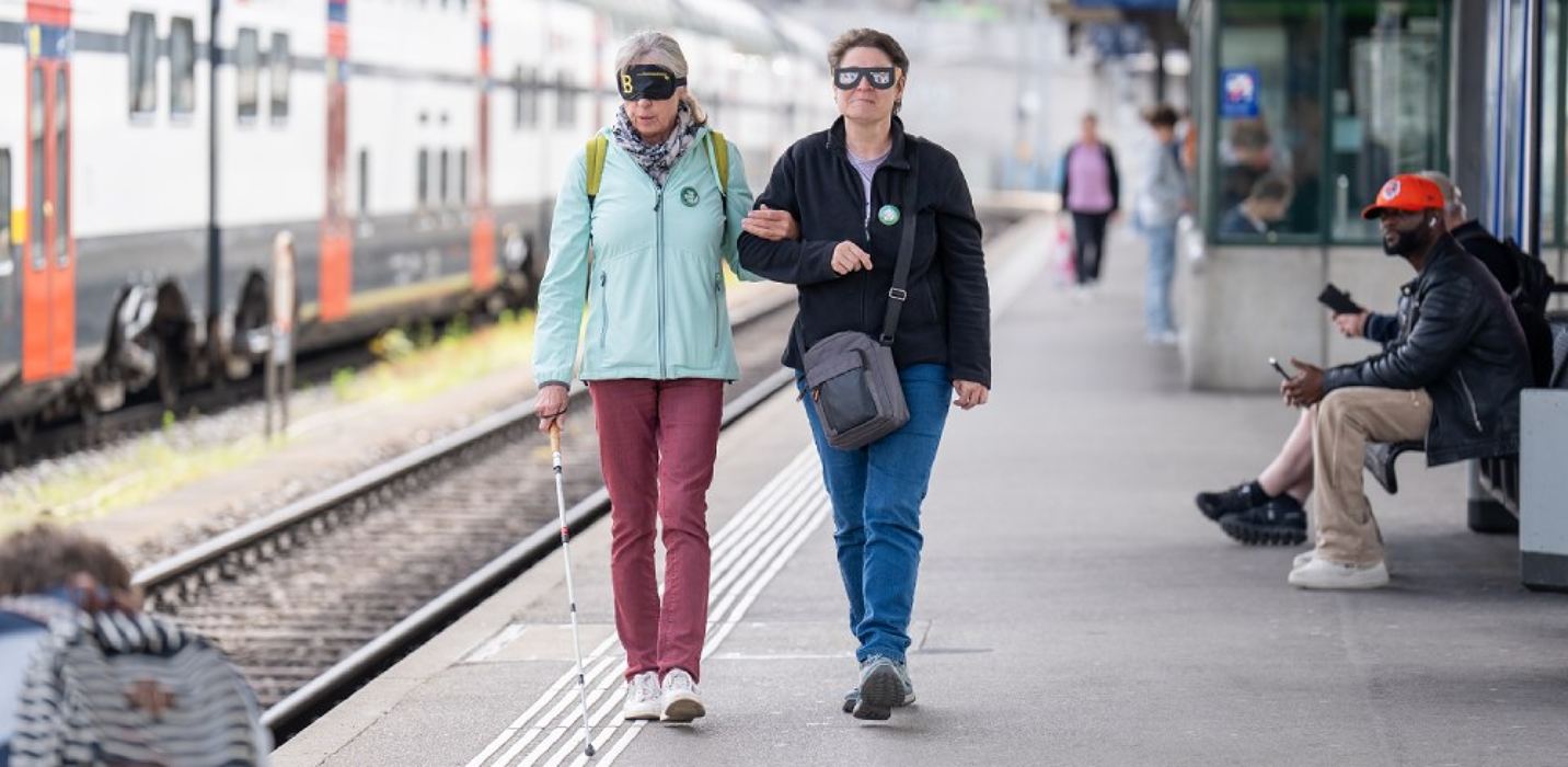 Zwei Personen laufen auf einem Bahnsteig entlang, eine hat einen Blindenstock Zwei Personen laufen auf einem Bahnsteig entlang, eine hat einen Blindenstock