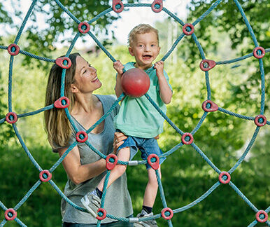 Kleiner blonder Junge mit Behinderung im Kletternetz auf Spielplatz, Mutter steht daneben. Kleiner blonder Junge mit Behinderung im Kletternetz auf Spielplatz, Mutter steht daneben.
