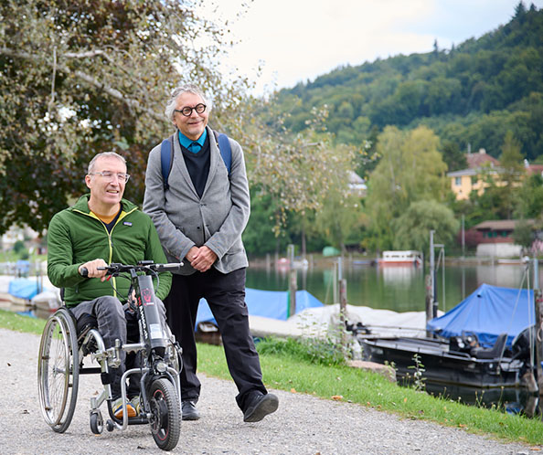Ein Mann im Rollstuhl fährt neben einem anderen Mann am Flussufer entlang.