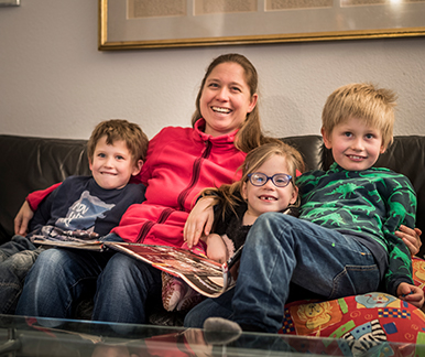 Mutter Sarah mit den 6-jährigen Zwillingen und dem älteren Bruder zu Hause auf dem Sofa. 